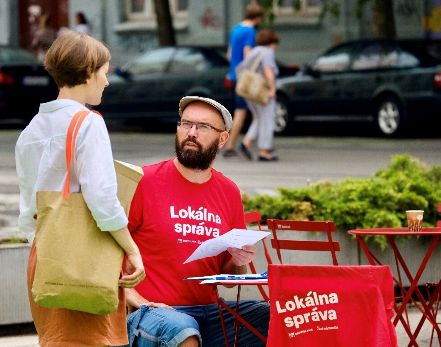 pán s červeným tričkom Lokálna správa sa rozpráva so ženou v bielej blúze a hnedej sukni, v pozadí sú ľudia prechádzajúci cez cestu