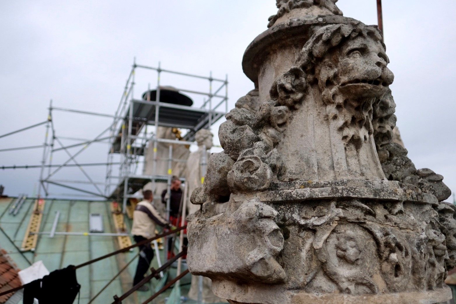 detail na jednu zo sôch na streche primácialného paláca, v pozadí sú robotníci na lešení, opravujúci inú sochu